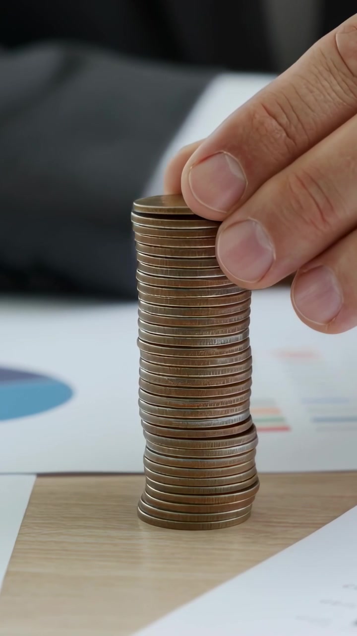 Vídeo vertical: Mão com manga de terno colocando moeda em pilha de moedas em mesa de madeira com gráficos