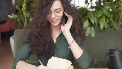 Young-businesswoman-talks-on-her-phone-and-writes-in-her-notebook-while-working-inside-a-café