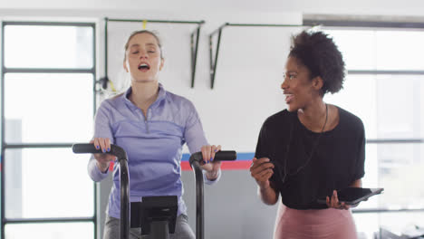 video de una entrenadora de fitness femenina diversa y una mujer en una bicicleta de ejercicio golpeando los puños en un gimnasio