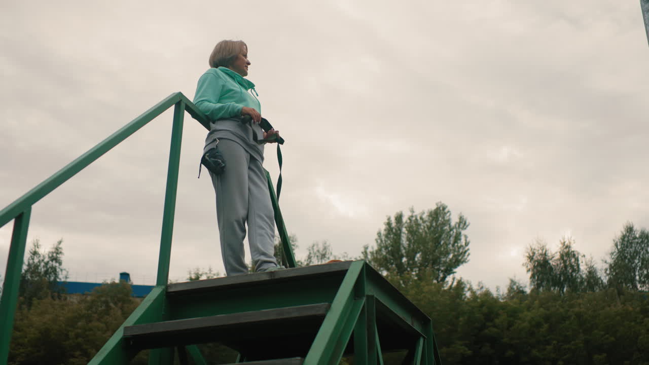 elegant lady standing confidently on high staircase in open park holding dog cord in hand looking around under cloudy sky surrounded by green trees relaxing after training session with dog companion