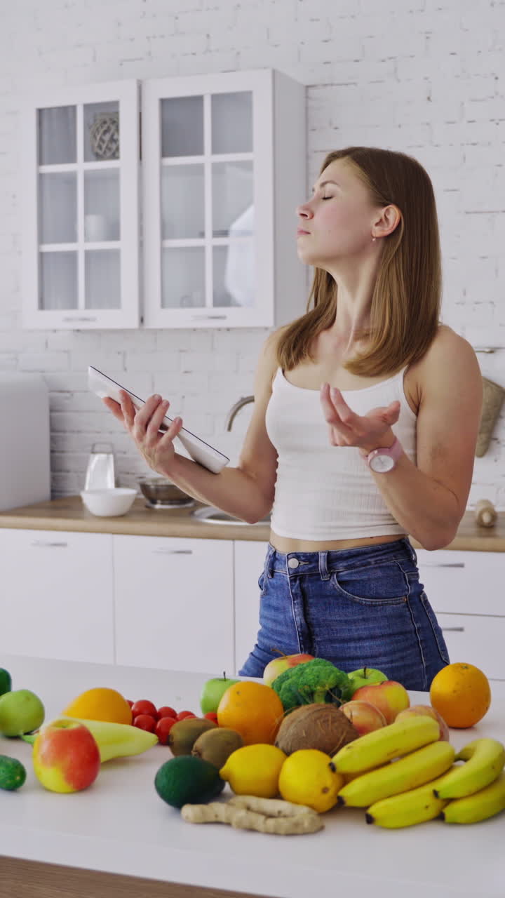 Pretty woman holds tablet in the kitchen. Young female is thinking about a new recipe of organic food. Fresh fruit and vegetables on the table. Healthy eating. Vertical video
