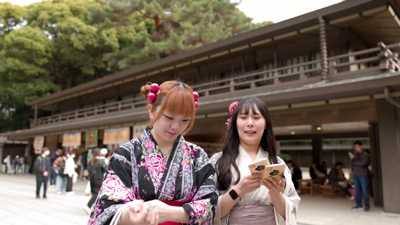 Due donne che indossano kimono tradizionali tengono in mano placche votive ema in legno mentre camminano attraverso un santuario giapponese.