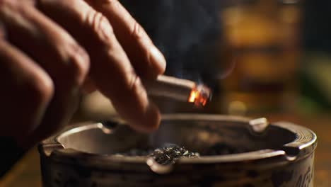 a hand delicately places a smoldering cigarette into an ashtray, showcasing the smoke swirling in the air, framed by a warm, intimate setting exuding a sense of relaxation and reflection