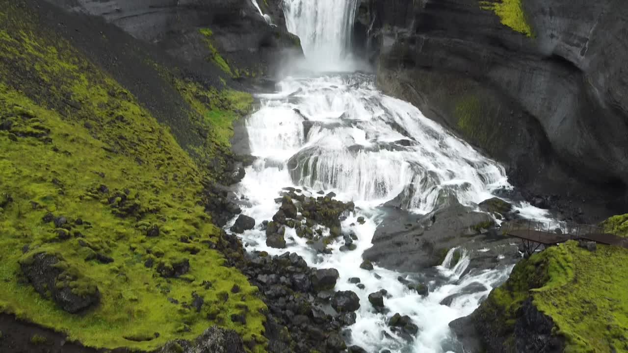 Drone shot of Mulagljufur canyon with tall waterfall plunging over dark cliffs surrounded by green moss in Iceland. Powerful flow carving through dramatic volcanic landscape in remote valley