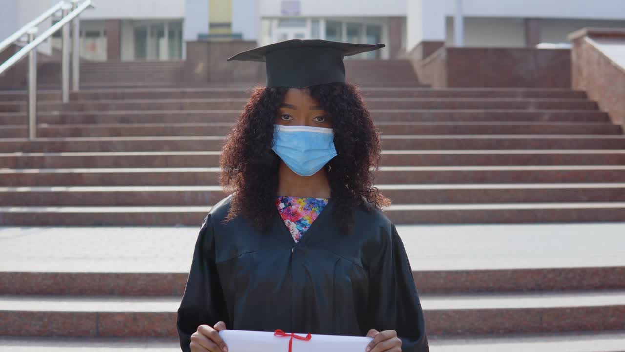 joven graduada afroamericana de pie frente a la cámara con un diploma en las manos. la estudiante tiene una máscara médica de protección. ella está de pie en las escaleras afuera.