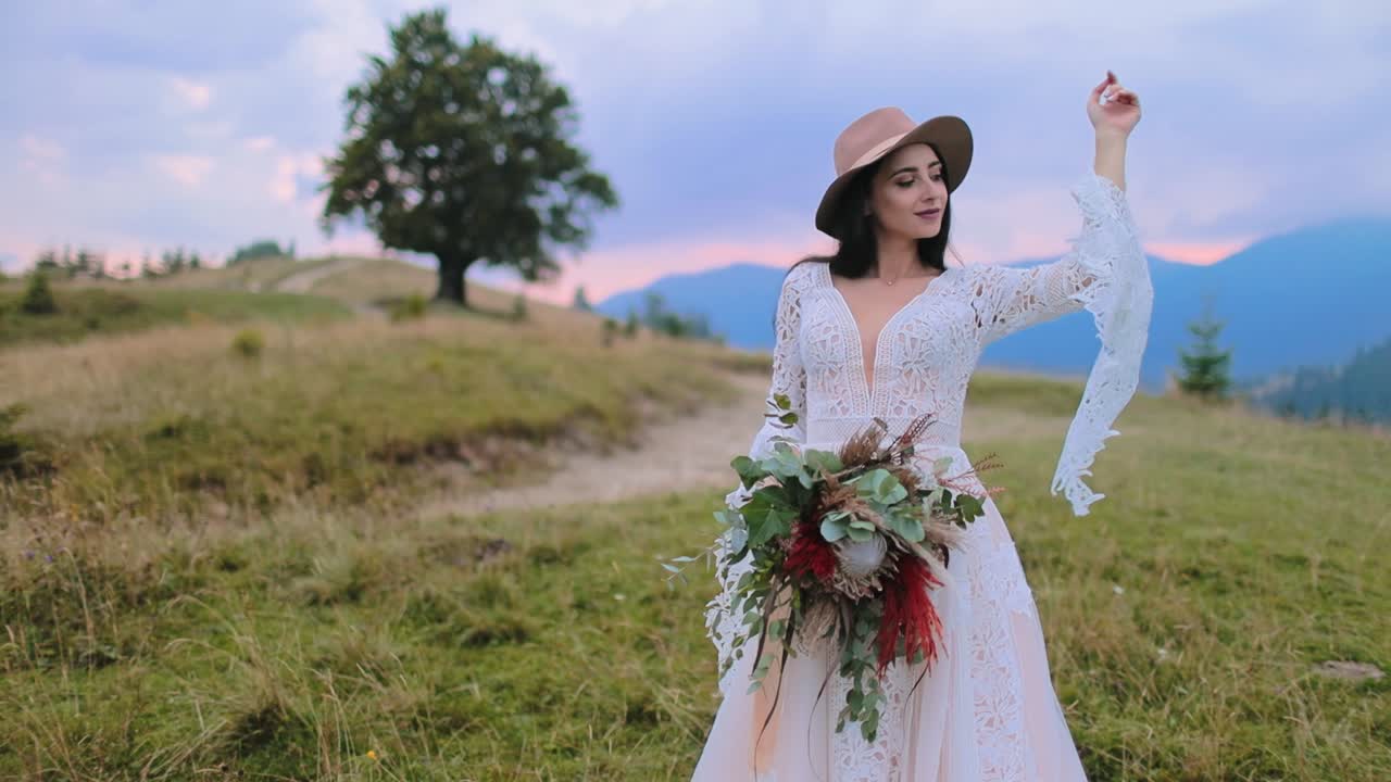 Beautiful bride among nature at sunset. Attractive woman in white wedding dress and in hat holding bouquet against the blue mountains.