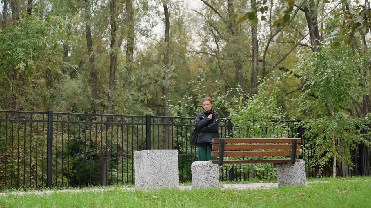 solitary person walking along park fence, autumn trees and wooden bench set reflective scene individual pauses, looks toward foliage, leans on stone block, breathes cool air, scattered leaves