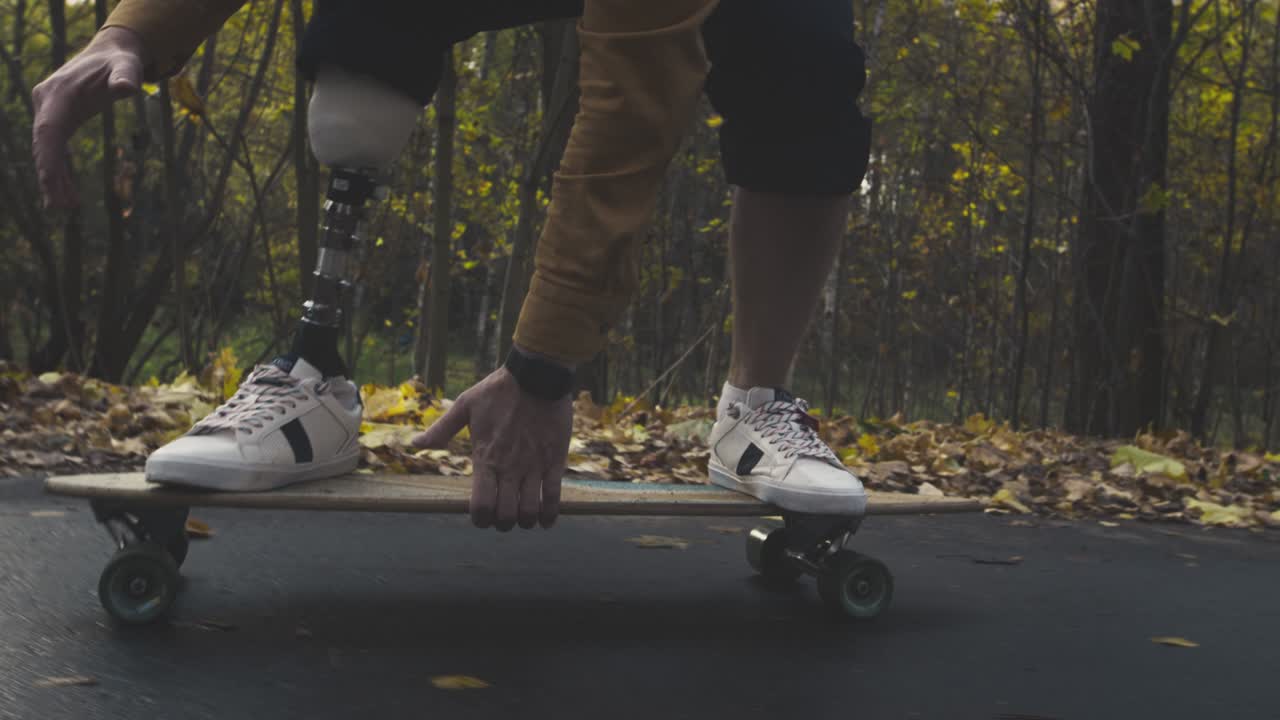 A young man with a metal bionic prosthetic leg is riding a skateboard in the autumn forest. An artificial leg pushes off the asphalt on a skateboard