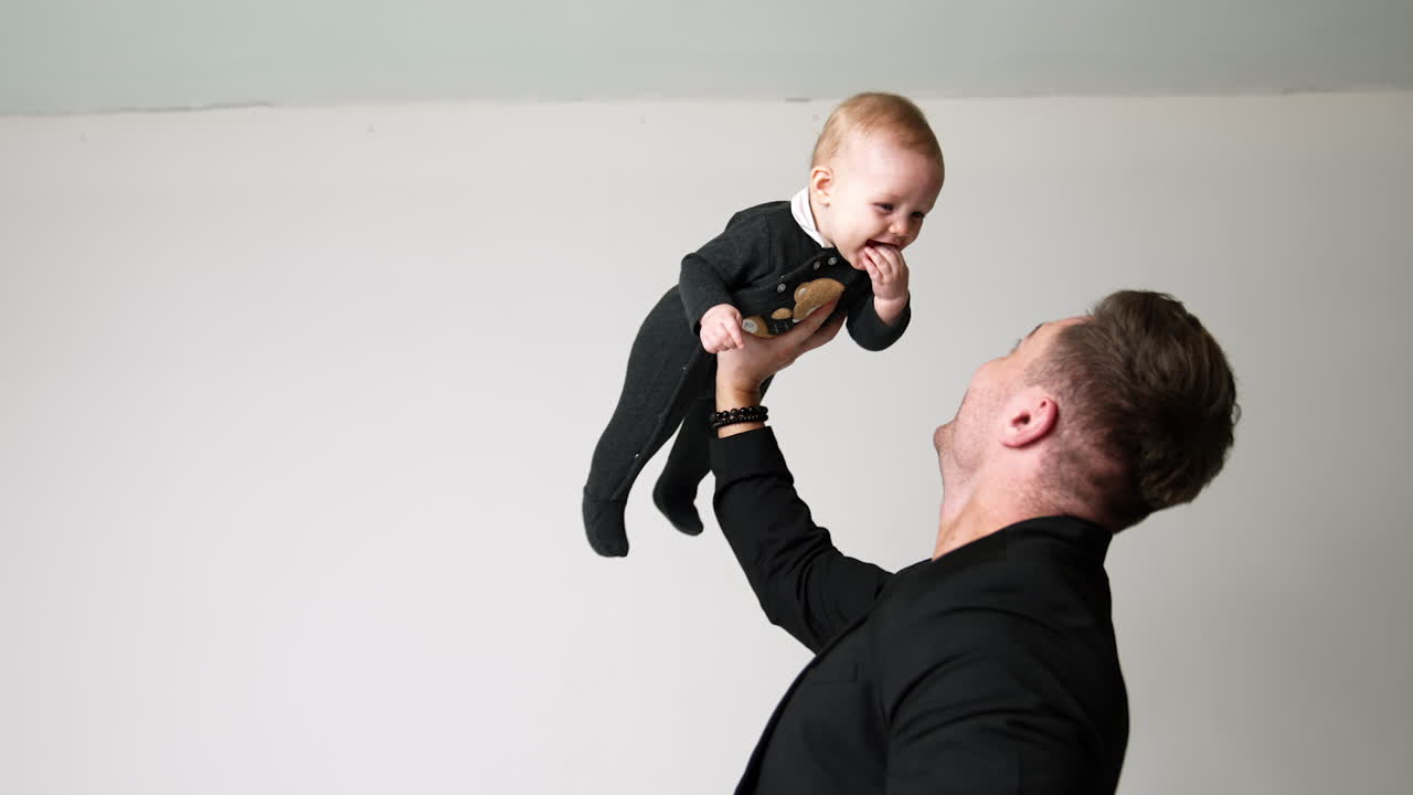 Strong Caucasian man in black jacket holding a baby up with one hand. Little baby keeps fingers in mouth and smiles to his father.