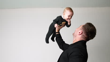 strong caucasian man in black jacket holding a baby up with one hand. little baby keeps fingers in mouth and smiles to his father.
