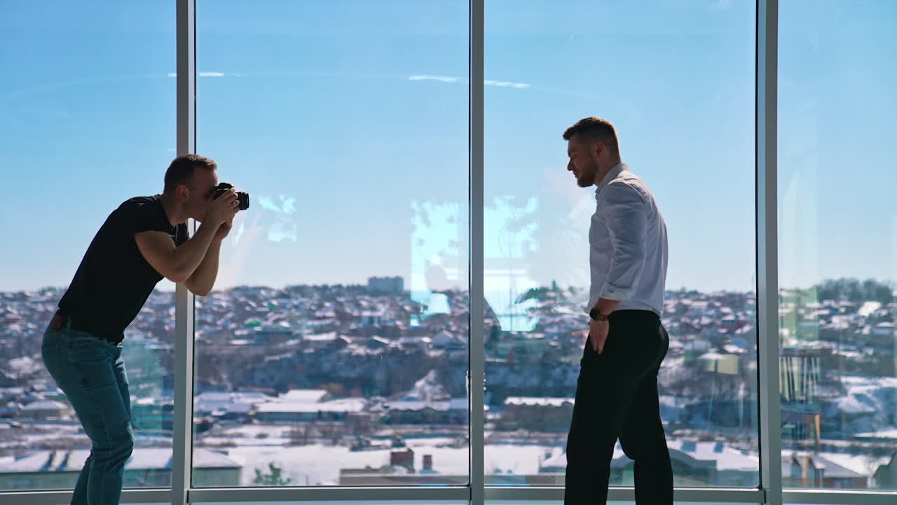 Handsome entrepreneur posing on camera. Young man photographing businessman near windows. Professional shooting in office. Profile view.