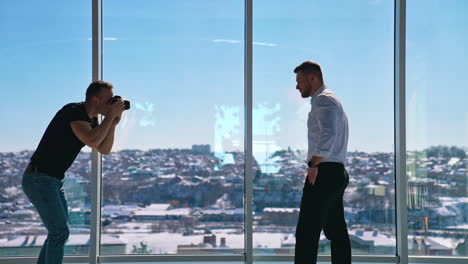 handsome entrepreneur posing on camera. young man photographing businessman near windows. professional shooting in office. profile view.