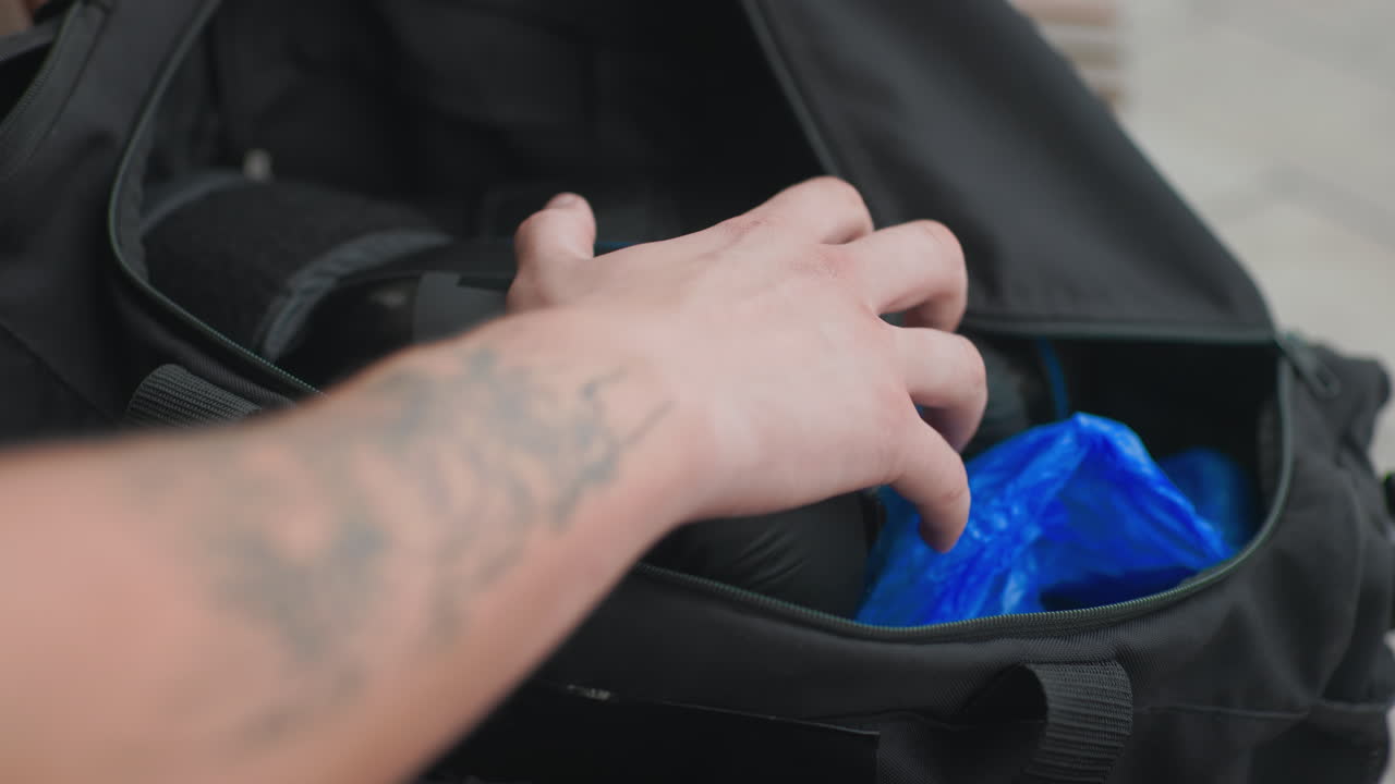 Una mano metiéndose en el bolsillo de una bolsa negra, revelando un vendaje de mano azul brillante y un guante acolchado, recuperación cuidadosa, brazo tatuado en movimiento, primer plano íntimo del compartimento interior, ambiente de práctica matutina