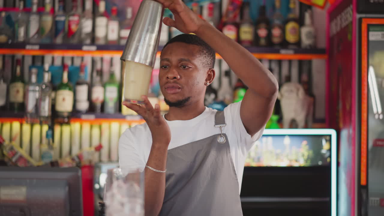 Focused bartender shakes cocktail in metal cup in bar. African American barman scrupulously prepares drink mixing ingredients in shaker. Black man works in bar