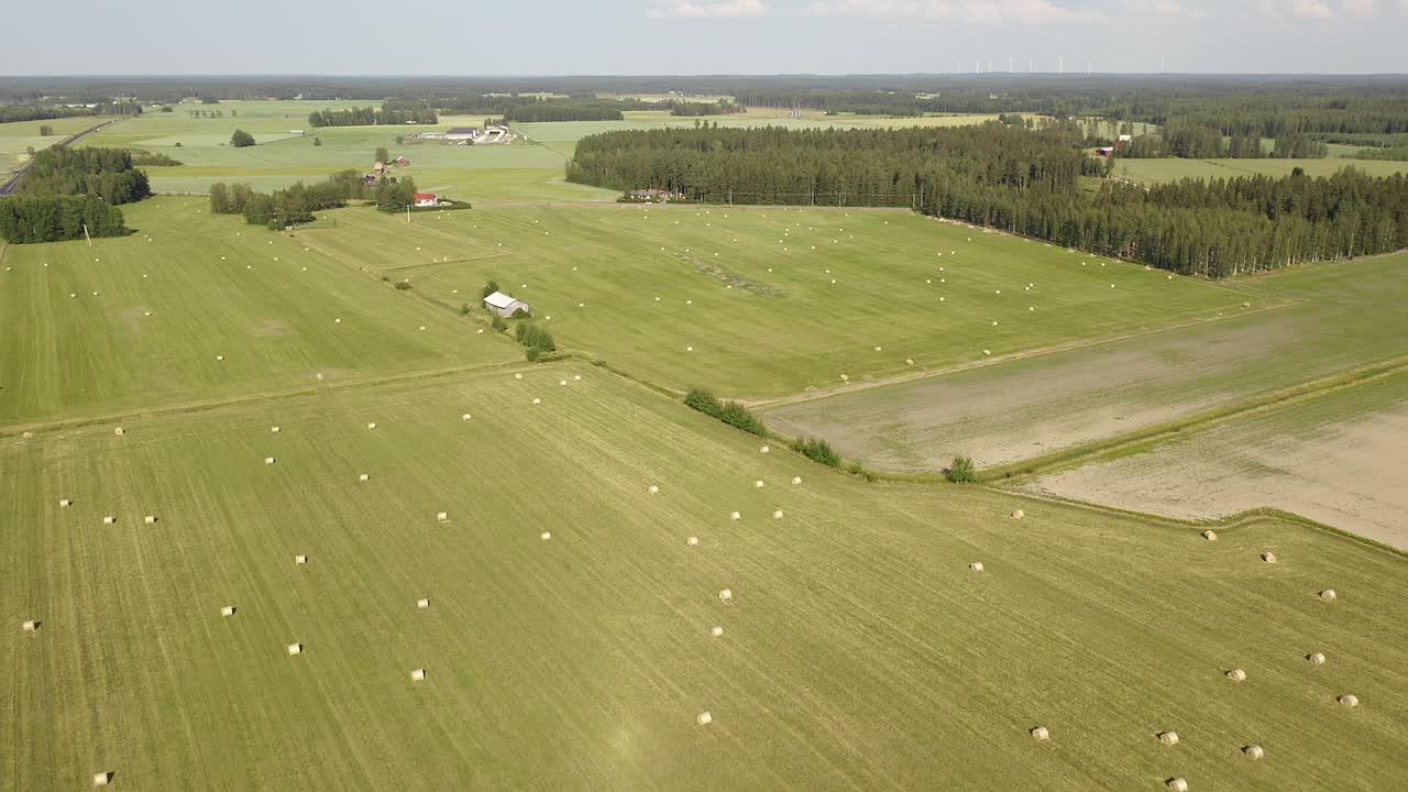 High aerial point of view of round haybales on a large grassy field with blue skies, scattered clouds and patches of forest in the background