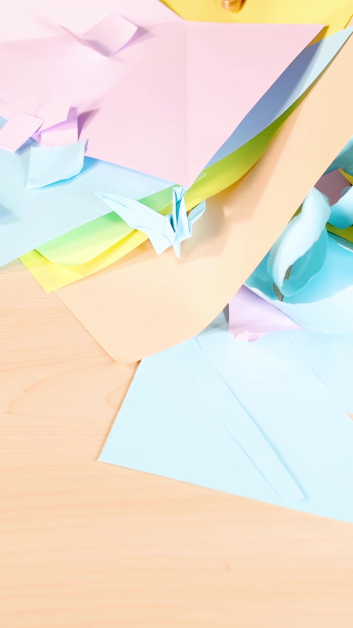 Hand chooses pastel origami paper on wooden desk, bright lighting, overhead static camera view