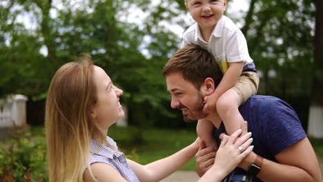 Happy-joyful-young-family-father,-mother-and-little-son-having-fun-outdoors,-playing-together-in-summer-park,-countryside.-Mom,-Dad-and-son-on-fathers-shoulders,-enjoying-nature-outside.-Slow-motion