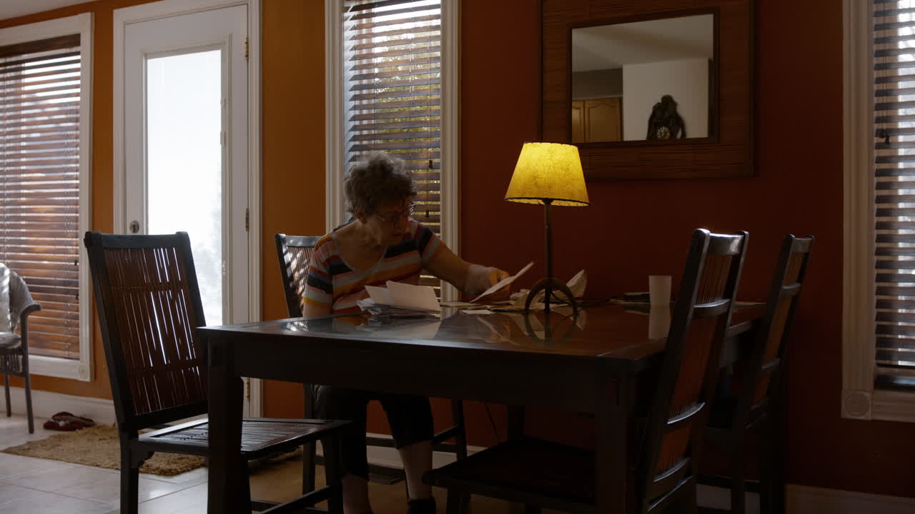 A senior lady sits alone at a dining room table, sorting through a pile of bills, and paperwork. Warmly lit, cozy home, by a table lamp. Orange painted walls, and brown furniture. Windy afternoon.