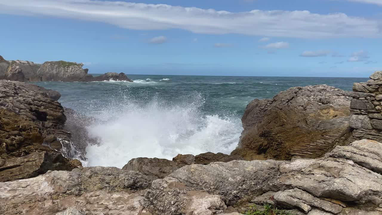 Ripresa al rallentatore di onde che si infrangono contro una costa rocciosa. L'acqua spumosa schizza sulle rocce mentre il mare blu scuro si muove sotto un cielo azzurro chiaro con una lunga nuvola