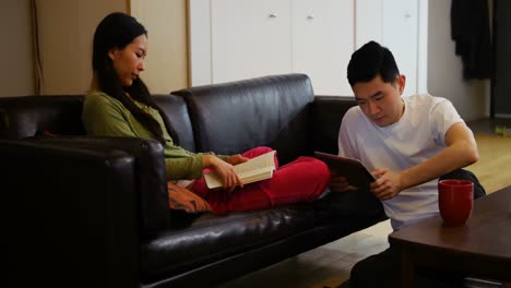 Woman-reading-book-while-man-using-digital-tablet-in-living-room