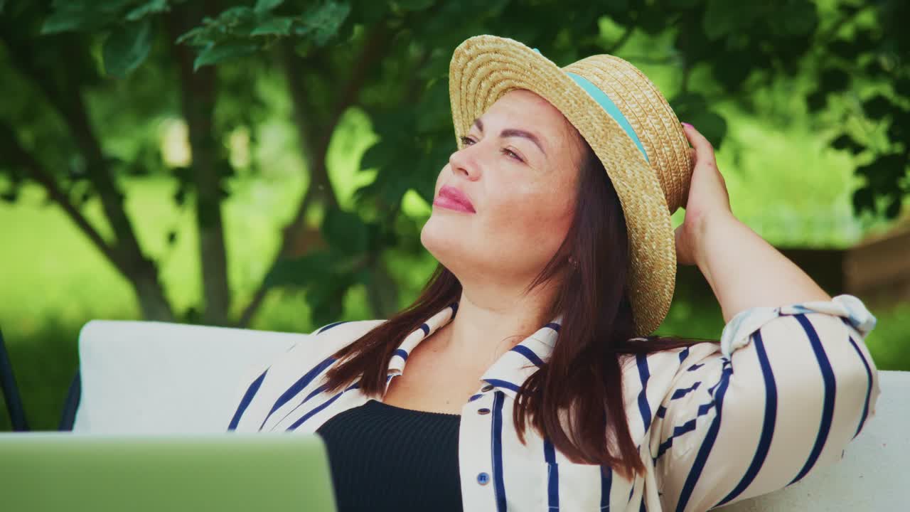 Woman relaxing outdoors in a straw hat, enjoying sunlight while sitting with a laptop