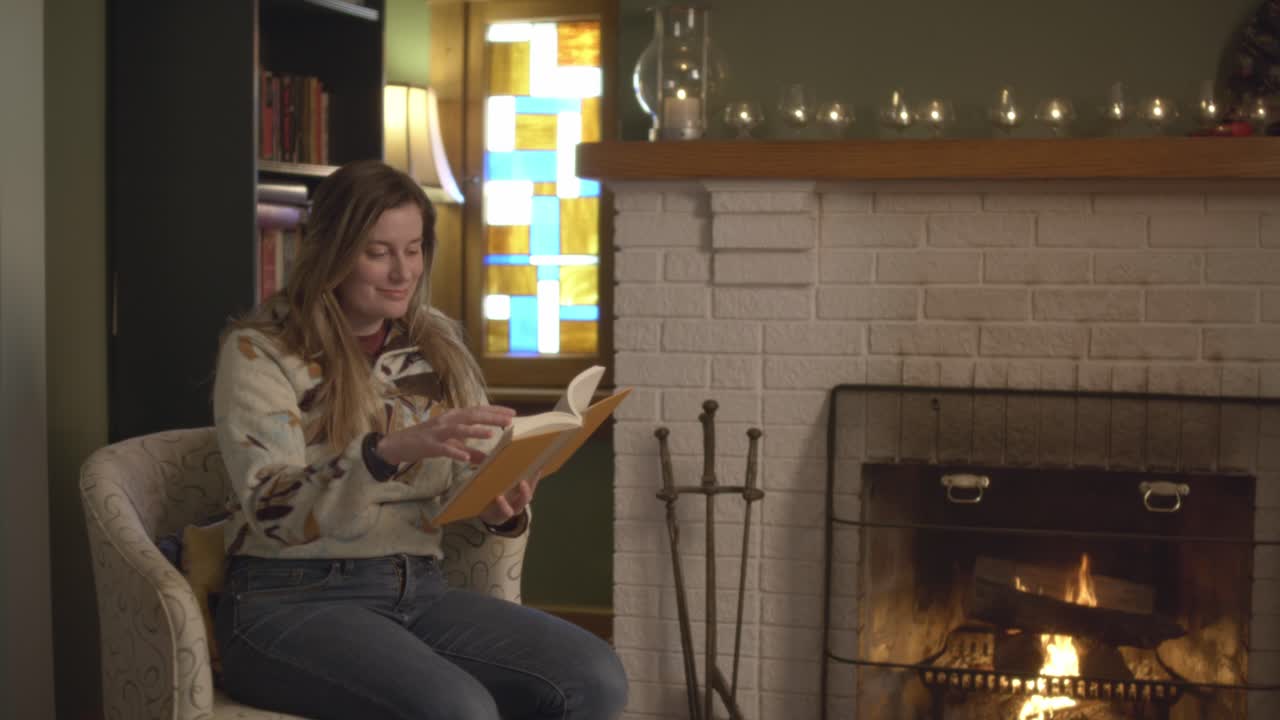 A woman in a sweater happily reading a book by a cozy brick fireplace. A warm fire in a relaxing fall setting with candles and a roaring log fire