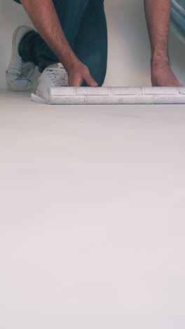 cu, dolly cam shot: happy young couple in workwear unrolls sheet of white wallpaper on floor in light renewing room closeup