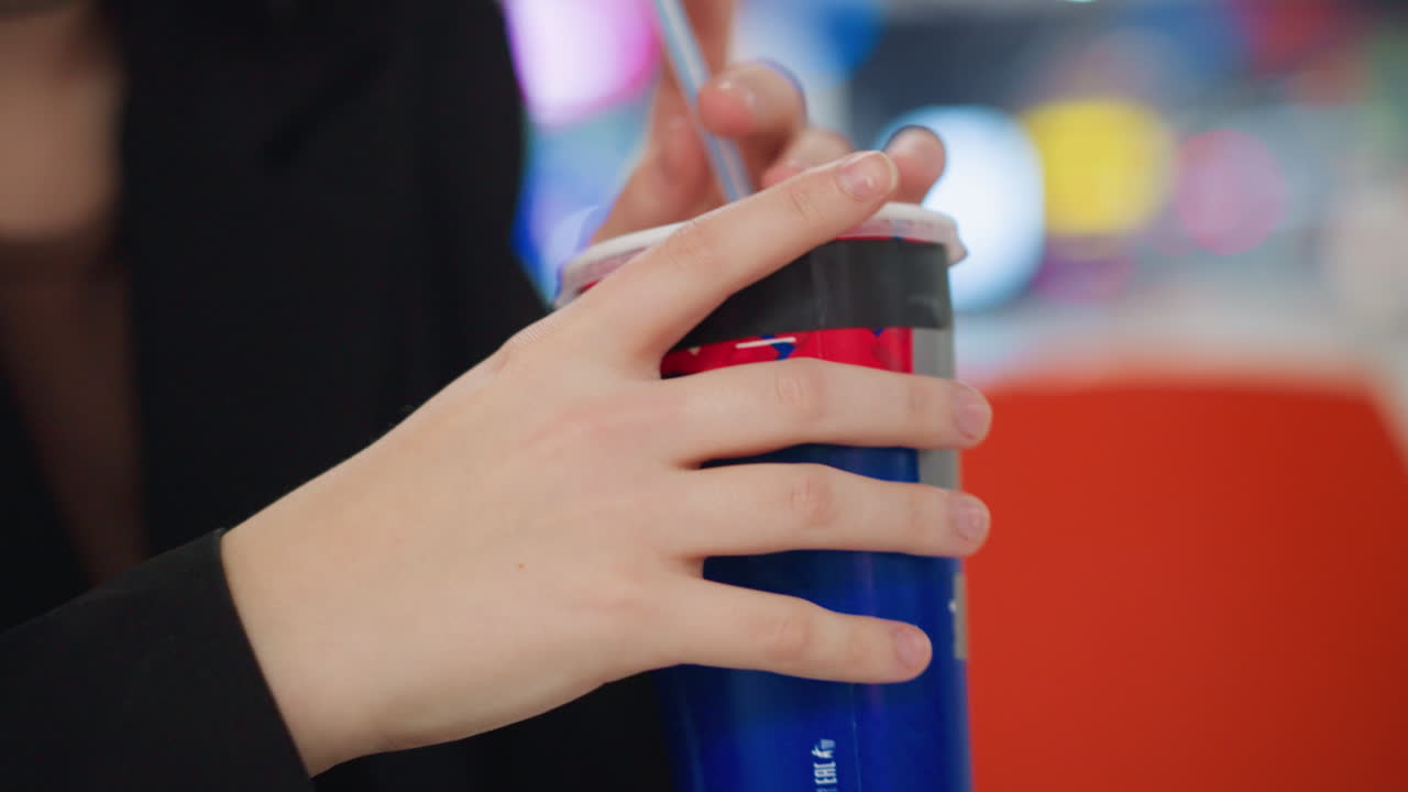Close up of young woman hand holding blue beverage cup with straw during casual moment, red and blue nail polish visible, blurred indoor background