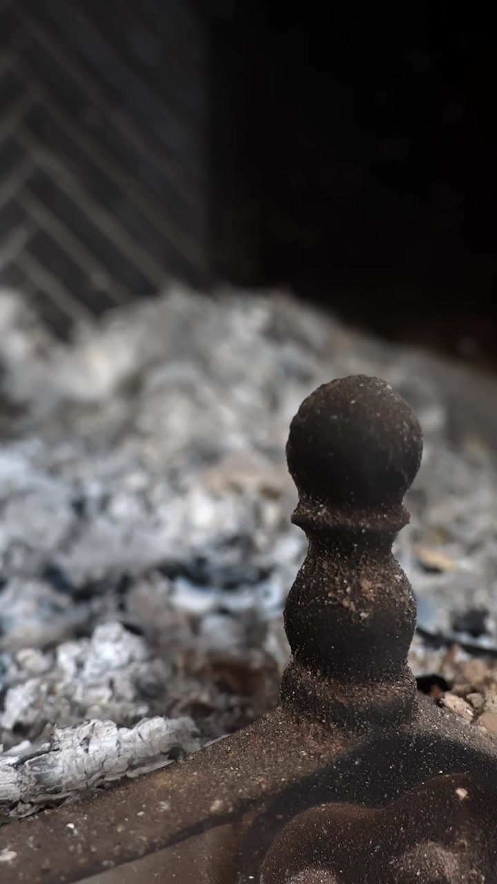 Vertical macro of cast-iron andiron finial beside gray ashes in a fireplace grate, shallow focus. Concept for home heating, soot cleanup, chimney maintenance, fire safety.