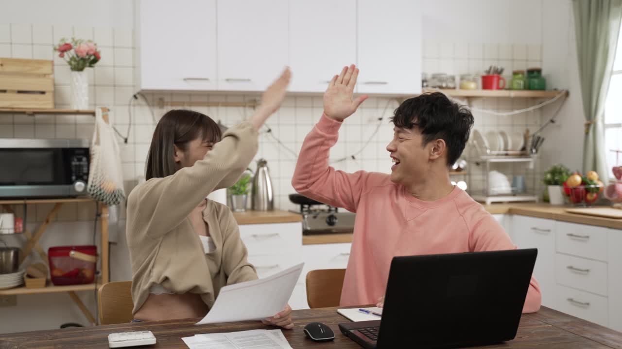 행복한 아시아 부부는 유리한 일자리 제안을 얻은 것을 기합니다. 아내는 남편이 온라인 킹을 사용하는 동안 박수를 치고 있습니다.