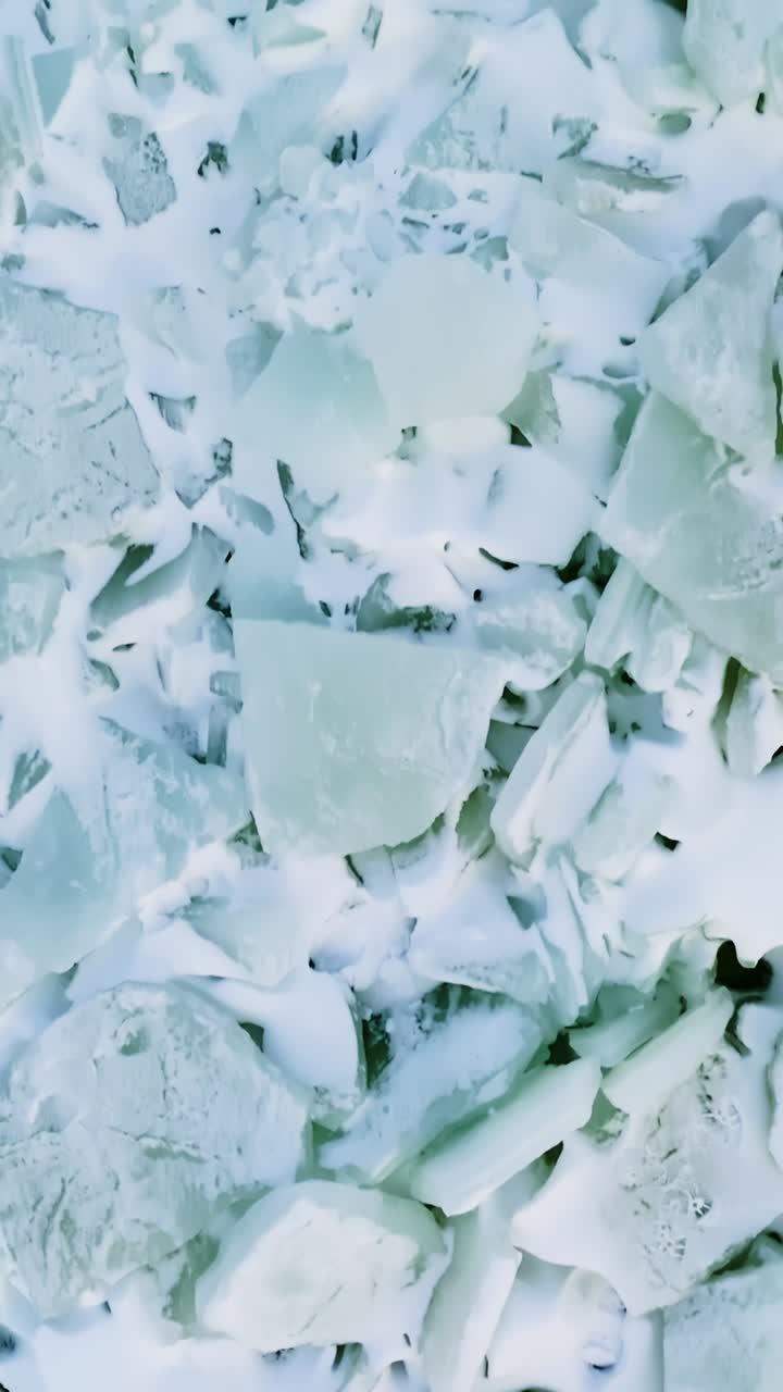 Primo piano verticale con drone di ghiaccio marino rotto e coperto di neve e tasche di fusione scure, creando texture invernali astratte e drammatici motivi polari su una superficie costiera ghiacciata.