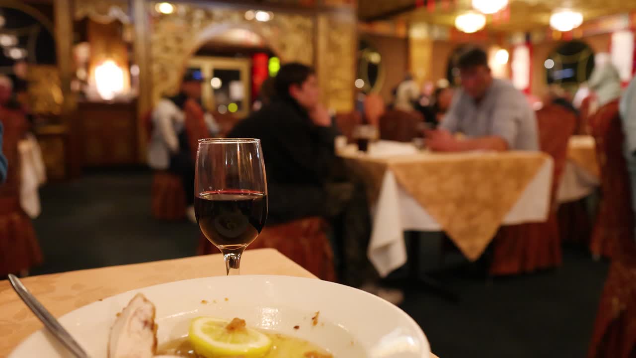 A glass of red wine sits on a table with a lemon dessert plate, surrounded by softly lit diners in a warmly decorated restaurant. Shallow depth of field and steady camera