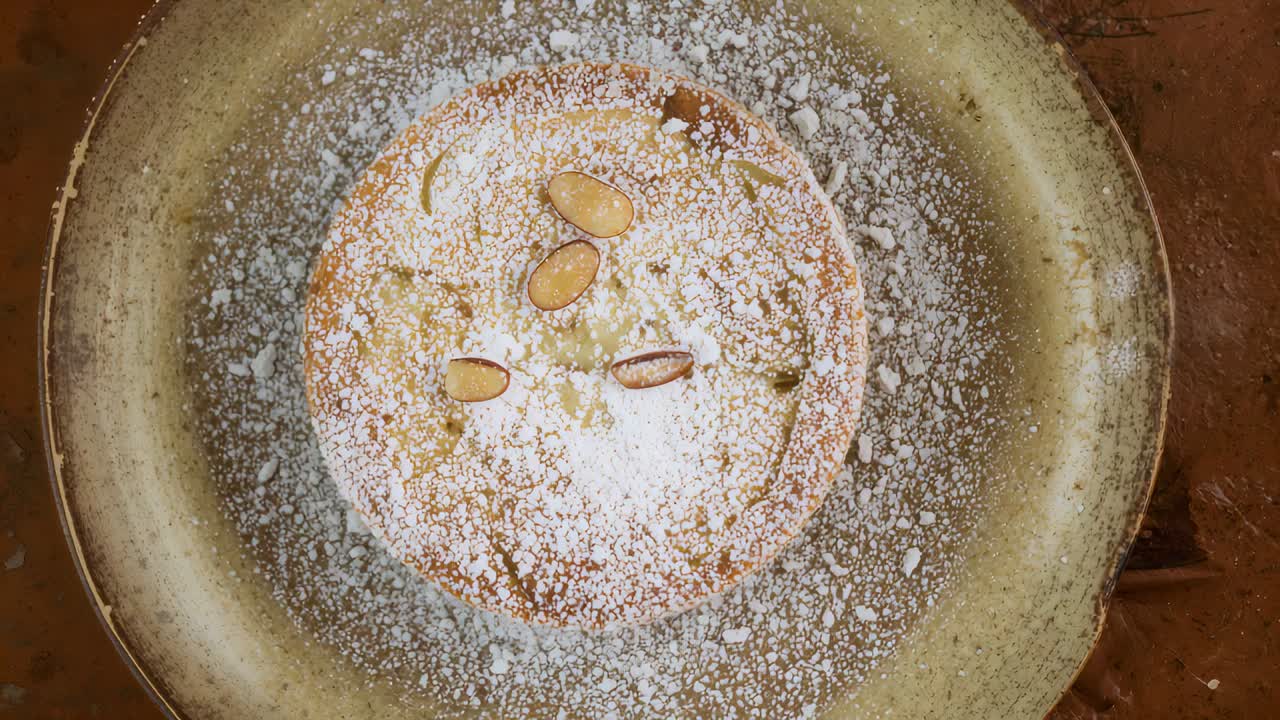 Moving camera reframing single round cake on wood surface, showing sugar dusting and almond slices