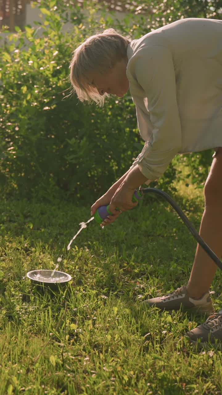 mujer usando una manguera para verter agua en una placa de metal en un terreno cubierto de hierba, con una segunda placa vacía cerca, la escena captura la luz solar natural y la vegetación con un edificio alto en el fondo