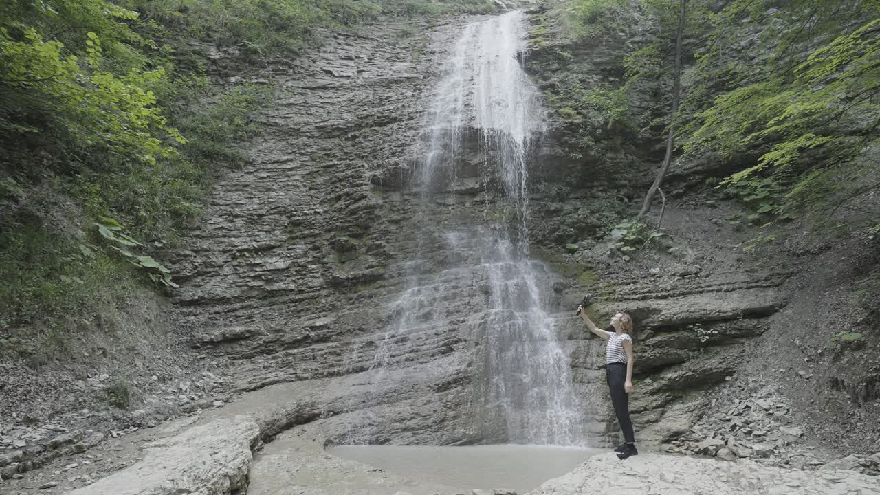 mujer tomando una foto de una cascada en un bosque de montaña