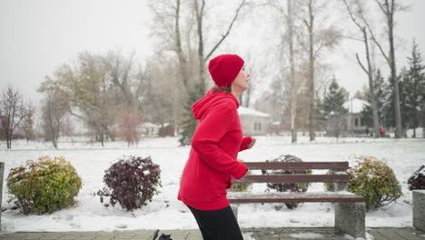 vista lateral de una mujer cansada corriendo por un sendero nevado en un parque con capucha roja y leggings negros, rodeada de bancos, arbustos coloridos, árboles helados de invierno y oficinas distantes cubertas de polvo de nieve