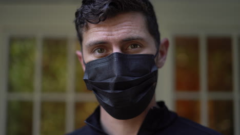 young man puts on a black face mask to protect himself and others from a virus then looks at the camera - 2020 global pandemic