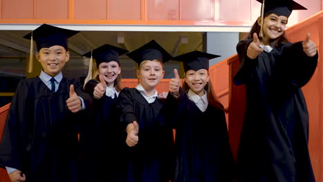 zoom in of happy kindergarten students in cap and gown looking at the camera and doing thumb up sign during the preschool graduation ceremony