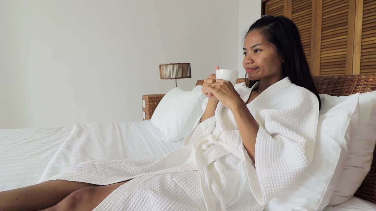 Giovane donna che indossa una vestaglia bianca e si sdraia su un letto d'hotel, bevendo caffè al mattino da una tazza bianca
