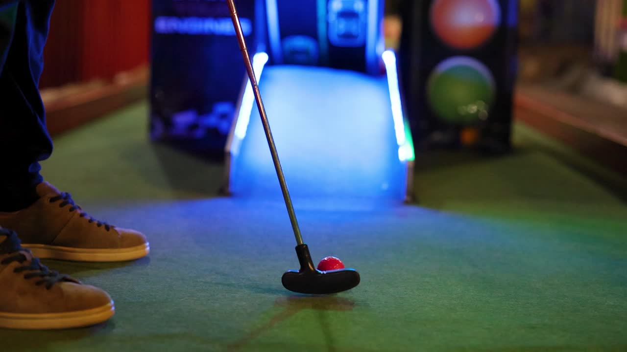A person attempts a mini golf putt under vibrant neon lighting, capturing the playful and focused atmosphere of the game