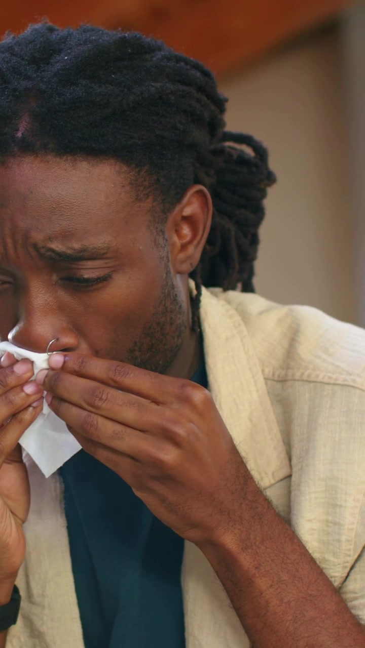 Vidéo verticale : Homme aux dreadlocks vêtu de sarcelle, utilisant un mouchoir blanc pour se moucher à la maison.