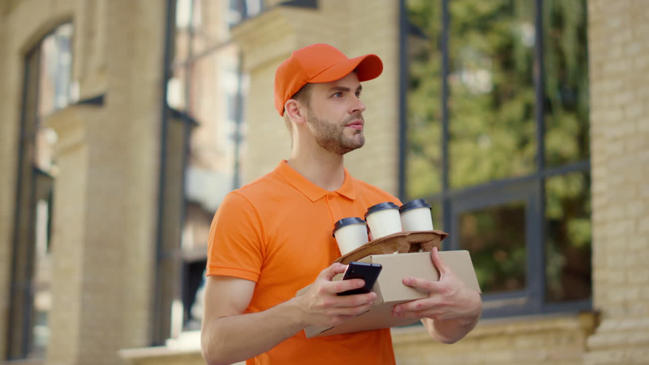 Courier calling mobile phone outdoor. Uniform guy delivering food and hot drinks