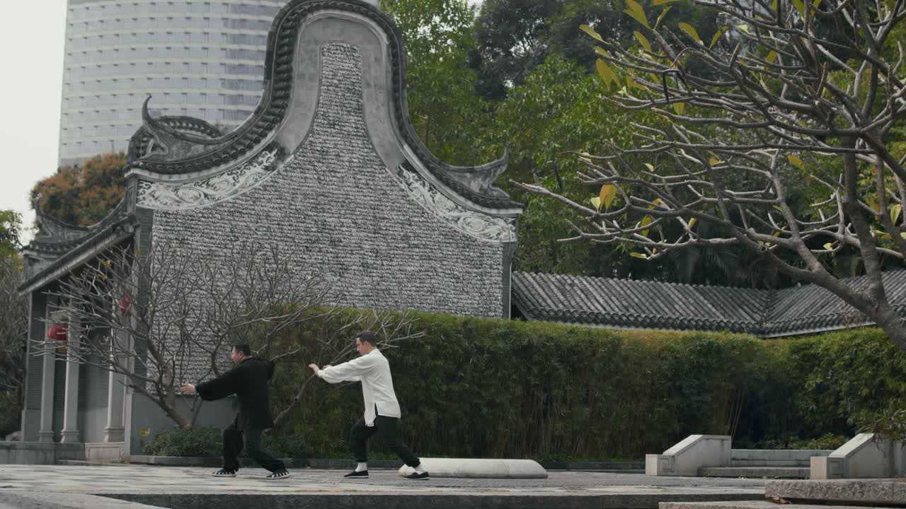 Two men practice Tai Chi in a traditional Chinese garden with a modern cityscape in the background