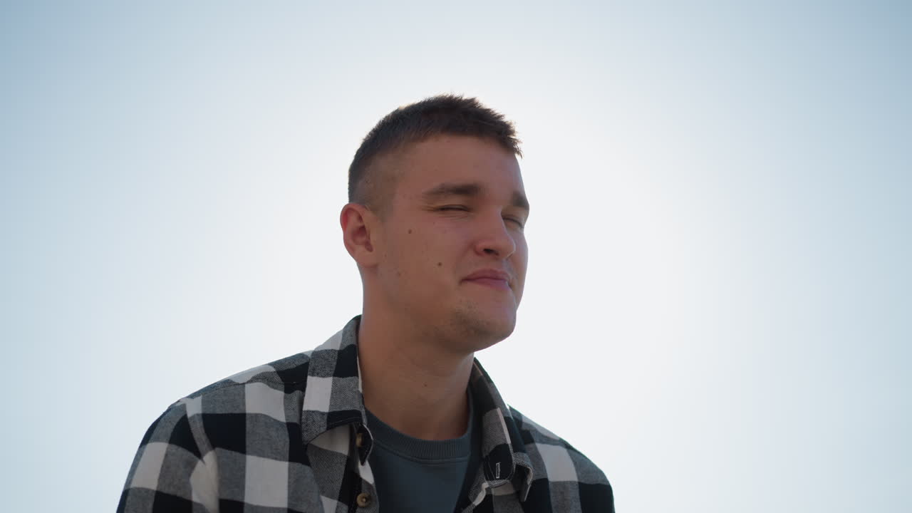 sunlight glows behind fair skinned man in checkered shirt as he gently closes eyes and puts something in mouth outdoors on bright day with serene blurred background