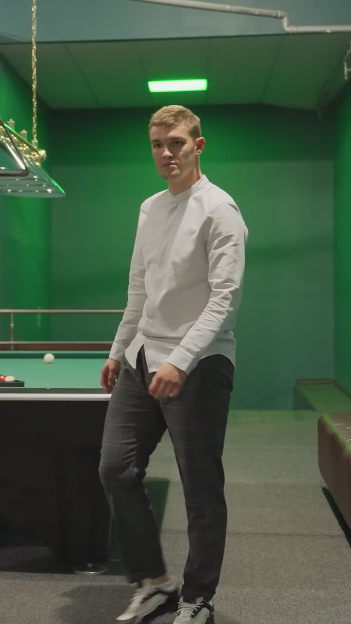 caballero con camisa blanca y pantalones grises entrando en la sala de billar al lado de la mesa de billar verde. elegante entorno interior con señales de billar, iluminación aérea, sofás de cuero y acento de pared de ladrillo