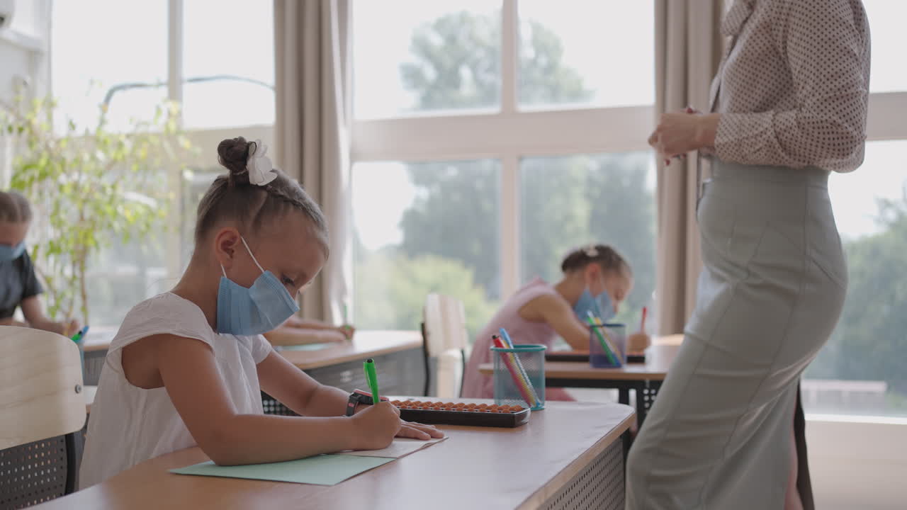 교실에서 선생님의 설명을 듣고 글을 쓰는 다민족 그룹의 아이들이 얼굴 마스크를 쓰고 수업에서 일하고 있습니다.