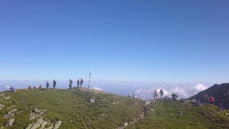 Los-Excursionistas-Se-Paran-En-La-Cima-De-Una-Montaña-Mirando-El-Paisaje-Con-Un-Cielo-Azul-Increíble-Y-Nubes-Bajas-En-El-Valle-De-Abajo