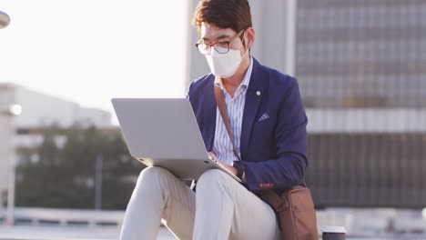 Asiatischer-Mann-Mit-Gesichtsmaske-Und-Laptop,-Während-Er-Auf-Der-Treppe-Im-Firmenpark-Sitzt