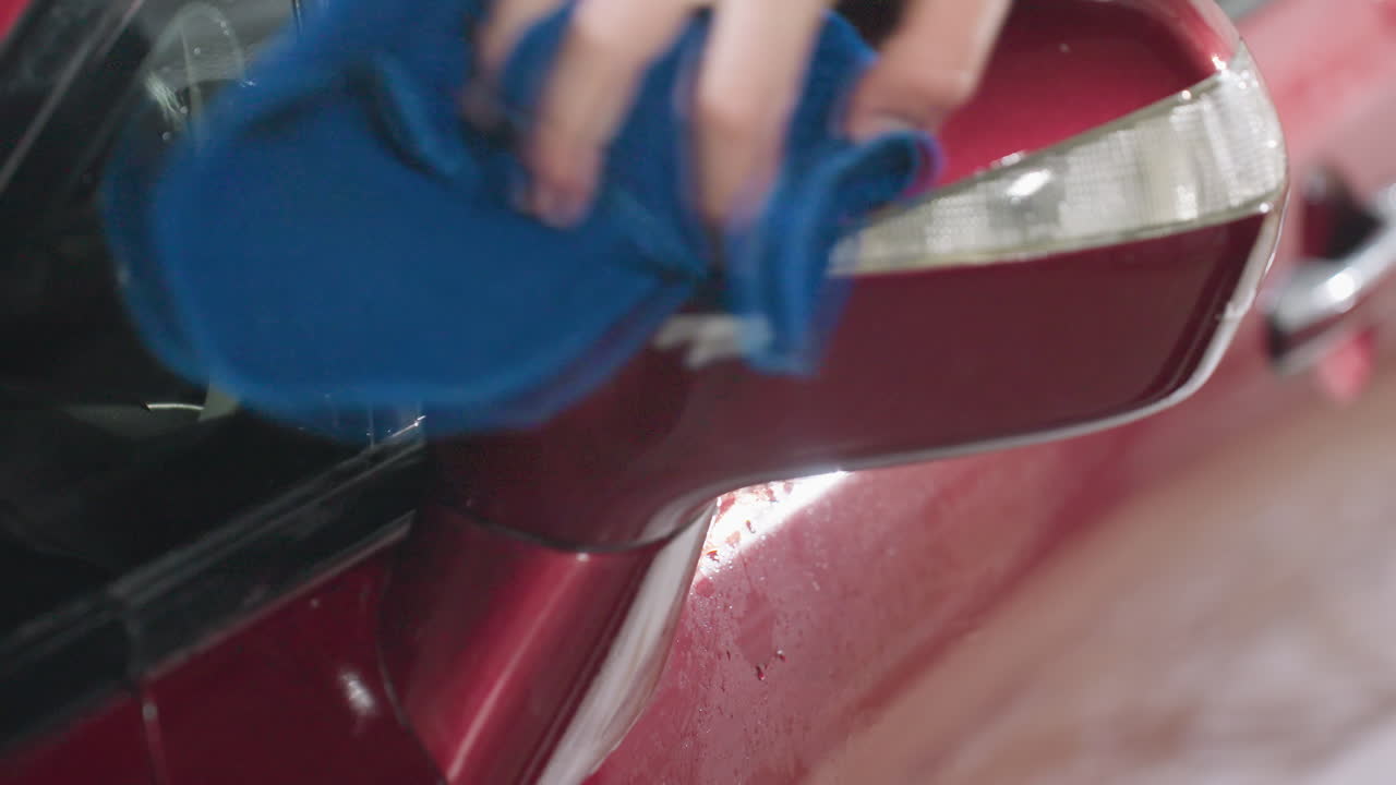 close up of red car side mirror being cleaned with blue rag by owner inside garage after washing with visible water droplets streaks and reflections under bright workshop lighting