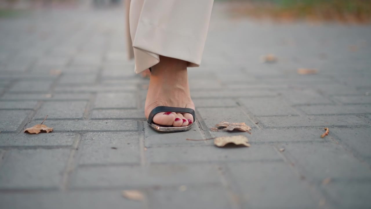 Set fra jorden ses fødder iført åbne sandaler med hæl, der bevæger sig over betonfliser, med den ene fod landende og den anden løftende i en jævn fremadgående bevægelse i blødt dagslys, med sandal, hæl